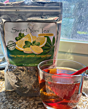 Load image into Gallery viewer, A sealed, silver bag of Sweet Yoni Tea which features a decorative label and a drawing of mint and lemons. The bag also features a large seevariety of herbs. In front of the bag is -through window to view the contents of the bag. Inside of the bag is aa glass mug that contains a tea infuser and an clear amber colored liquid. The tea bag and mug are in front of a sunny window on a granite countertop.
